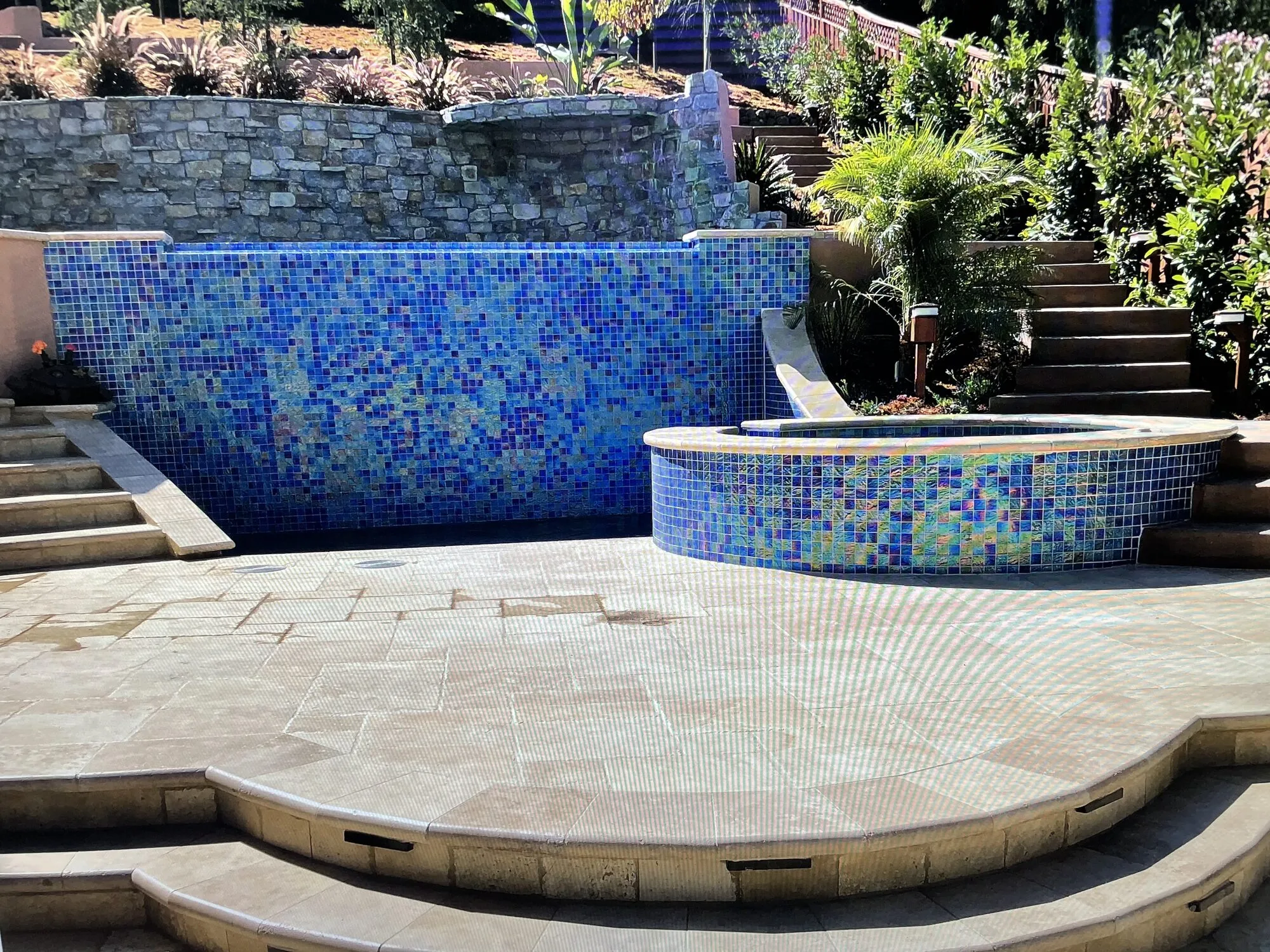 A circular mosaic-tiled spa and deck framed by a stone retaining wall and stairs to the next level.